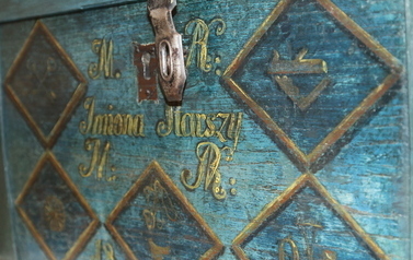 Wystawa stała 'Historia Wronek (od) nowa' - fragment malowanej skrzyni drewnianej z okuciem z XIX wieku. Skrzynia jest pomalowana w kolorze niebieskim, ozdobiona rombami w kolorze złotym oraz złotymi inicjałami.