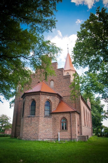 Ceglana fasada poewangelickiego kościoła w Wartosławiu. Dookoła dużo zieleni, trawnik i drzewa liściaste. 