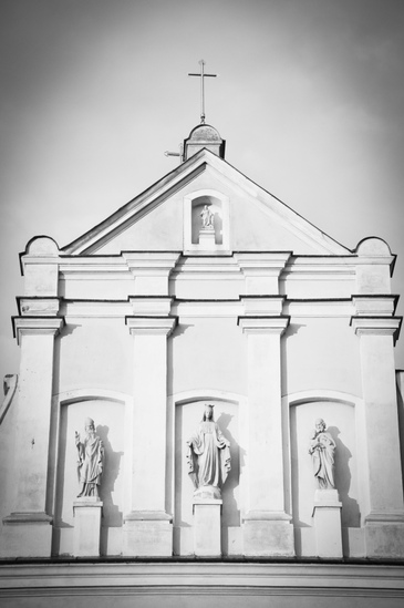 Fragment fasady kościoła braci Franciszkanów. Zdjęcie w odcieniach szarości. Widoczna sygnaturka z krzyżem oraz trzy rzeźby. 