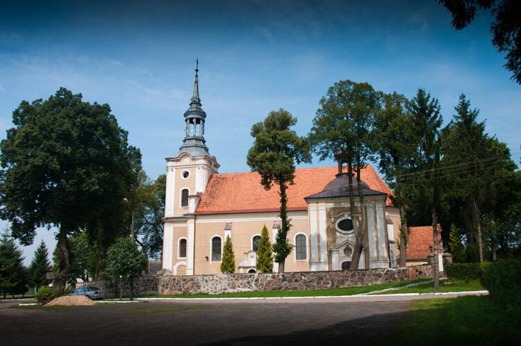 Kościół świętego Krzyża i Mikołaja w Biezdrowie. Widoczny kościół z fasadą w kolorze beżowo-pomarańcowym oraz dach pokryty czerwoną dachówką. Obok rosną rozłożyste drzewa liściste. 